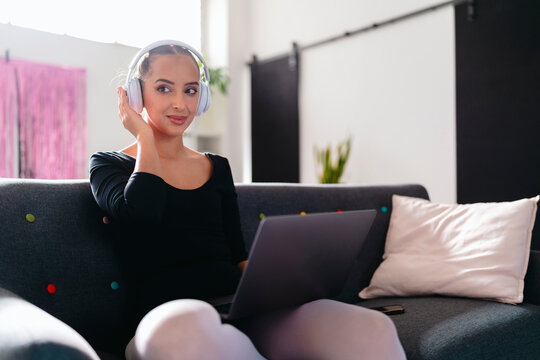 A young ballerina sits on a gray couch, wearing white headphones and interacting with a laptop, in a bright modern studio featuring pink curtains and vibrant decor.