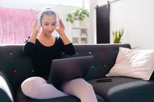 A young ballerina sits on a gray couch, wearing white headphones and interacting with a laptop, in a bright modern studio featuring pink curtains and vibrant decor.