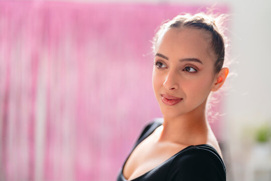 graceful ballerina poses with arms delicately extended upward in a bright studio with pink accents, her black leotard and white tights creating a striking visual contrast.