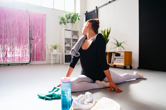 A ballet dancer wearing a black leotard and white tights stretches on the floor of a bright studio with pink decor and indoor plants creating a lively atmosphere.