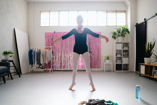A ballerina in a black leotard and white tights prepares for a pli&eacute; in a spacious studio, with bright natural light streaming through the windows and a pink shimmering curtain in the background.