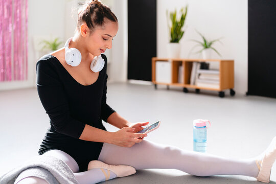 young woman wearing a black leotard and white tights sits on a dance studio floor with her legs stretched out, holding a smartphone while wearing headphones around her neck