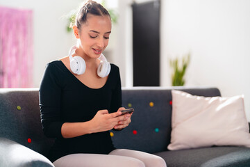 A close-up of a young ballet dancer with white headphones around her neck, smiling while using her phone, dressed in black dancewear with white tights in a bright indoor room.