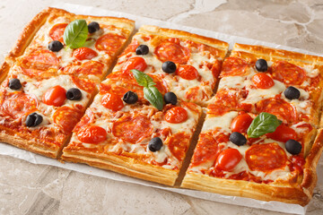 Tasty baked Puff pastry tart pizza style with pepperoni sausage, olives, tomatoes and mozzarella cheese close-up on parchment on table. Horizontal