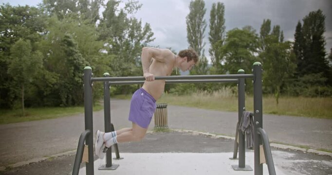 A young man performs dips on parallel bars in a park. Two friends stand by and give him encouragement.