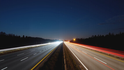 Fototapeta premium A long exposure shot of a highway at night, with light trails on the road, grass on the ground, trees in the background, and a sky with clouds.Wallpaper, banner.