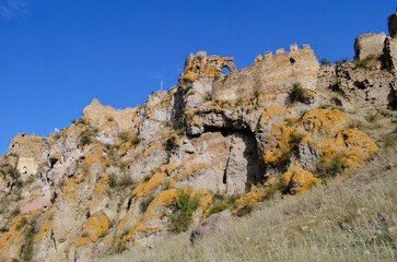 A stunning view of ancient stone ruins perched on a rocky hillside, partially covered with vibrant yellow moss, under a vivid blue sky. The crumbling stone walls and arched openings add a sense of his