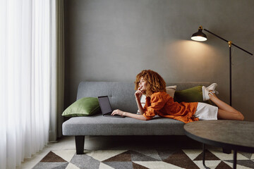Young woman working on a laptop in a modern living room with green accents, showcasing a relaxed...