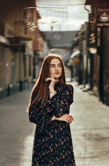 girl with long red hair standing on the street. She is wearing a dark dress with a floral pattern.