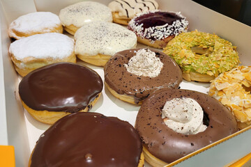 A box of assorted gourmet donuts featuring a variety of toppings and fillings, including chocolate glaze, powdered sugar, vanilla frosting, crushed nuts, and whipped cream.