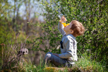 A little boy sits on the grass and greedily drinks orange juice