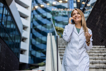 Fototapeta premium A cheerful female nurse smiles as she talks on her smartphone as she walks to work in a city medical building. After work. Nurse using her mobile phone while taking a break.