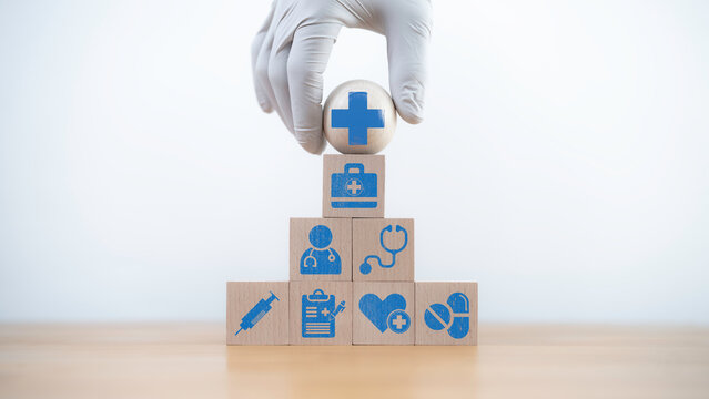 Hand in a medical glove carefully places a wooden health block atop a pyramid of blocks with medical icons. Symbolizing healthcare growth, responsibility, and safety