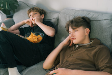 Two tired teenager brothers lying on sofa looking bored, watching tv, eating snacks at home....