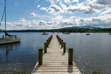 Fototapeta premium View over length of small boat pier into Lake Windemere in Lake District National Park near Ambleside, UK with sailboats in water against a white clouded blue sky