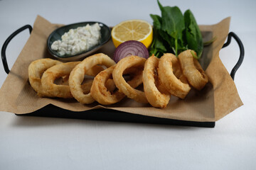 crispy fried calamari rings , golden brown crusty finger food served with dip sauce, greens and lemon, styled on plate with cooking paper