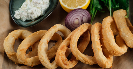 crispy fried calamari rings , golden brown crusty finger food served with dip sauce, greens and lemon, styled on plate with cooking paper
