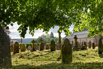 Naklejka premium Graveyard with headstones of the St Michael the Archangel Church in Chagford, UK seen from under lush green trees and early morning sun shining through