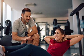 Happy athletic woman doing sit-ups while having sports training with personal coach in gym.