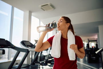 Young sportswoman drinking water from bottle while exercising in fitness center.