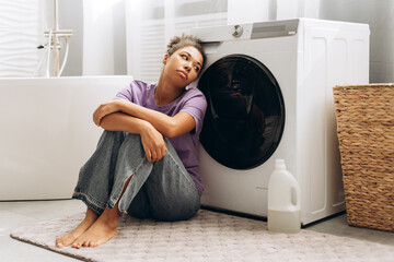 Exhausted housewife leaning on washing machine in bathroom
