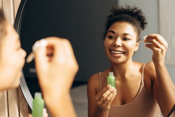 Smiling woman applying face serum with pipette in bathroom mirror