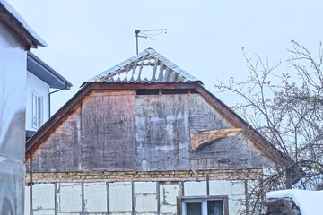 part of an old rural house with a gray wooden attic made of plywood under a slate roof in white snow against the sky on a winter street