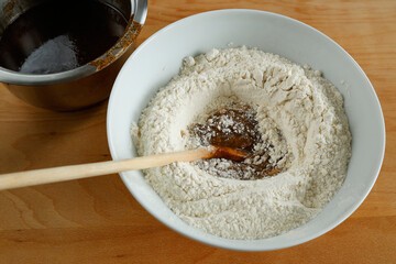 Making gingerbread. A bowl of flour and dry ingredients is being mixed with a buttery, spiced liquid from a saucepan. A wooden spoon rests in the bowl for stirring.