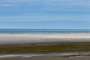 Calm ocean waters meet a wide, light-colored beach.  A small ship is visible on the horizon.  Low light and muted colors.
