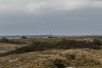 Coastal dunes, a light-gray sky, and a distant red-and-white lighthouse.  People are cycling on a path.  Cloudy day.