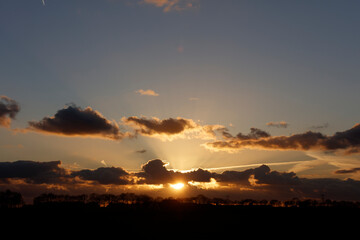Dramatic sunset through a layered sky of clouds.  Silhouette of trees and a landscape line against the warm hues of the setting sun.