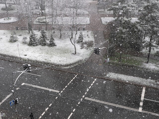 View from the window over heavy fluffy snow falling over the pedestrian crossing with red traffic light and urban park with a lonely person walking. Snowy winter day.