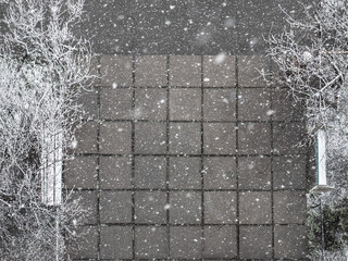 Large fluffy snowflakes falling over square tiles, trees, benches at the building entrance. Snowy winter day.