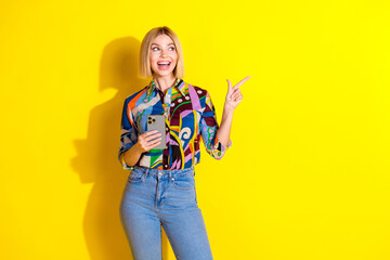 Photo of attractive young woman hold gadget point look empty space dressed stylish colorful clothes isolated on yellow color background