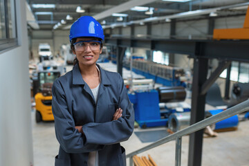 Confident young engineer, an Indian woman, in a blue helmet and gray work coat at a factory, portrait shot.