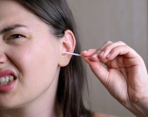 Millennial woman cleaning her ears in the bathroom, feeling pain during ear cleaning and making a grimacing facial expression.