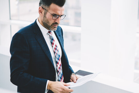 Confident thoughtful proude ceo in formal wear analyzing information data of accounting reports tanding in office.Pondering mature entrepreneur 50 years old thinking on banking text documentation
