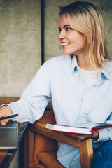 Happy blonde freelancer working remotely at digital netbook device connected to wireless 4G internet.Positive hipster blogger with textbook in hand laughing during distance job at laptop computer