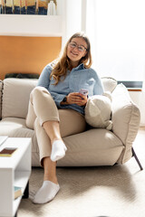 Beautiful middle aged woman sitting on sofa, holding mobile phone, looking at camera