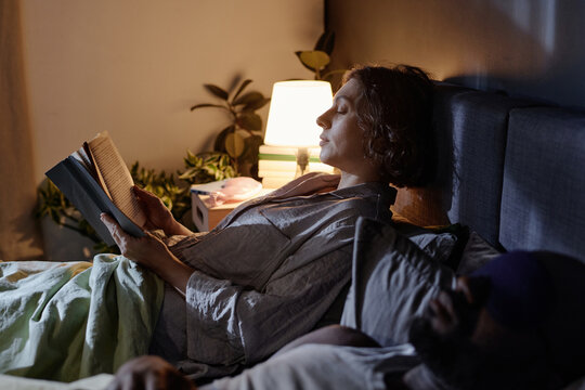 Medium close up of woman in cotton pajamas resting in bed and reading novel while her husband sleeping next to her - Powered by Adobe