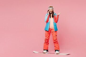 Full body young woman wear warm blue jacket ski goggles mask spend weekend winter season in mountains riding snowboard talk speak on mobile cell phone isolated on plain pink background. Hobby concept. © ViDi Studio