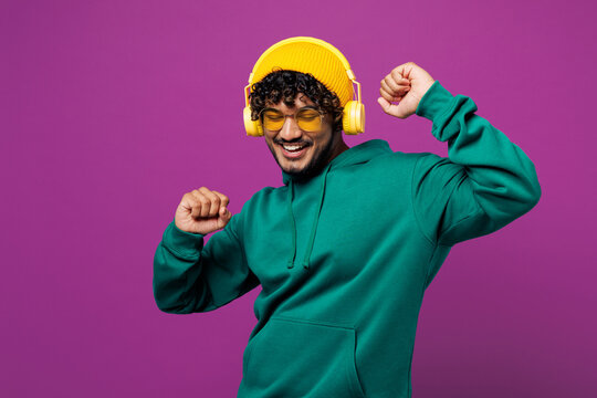 Young happy Indian man wear green hoody yellow hat sunglasses casual clothes listen to music in headphones raise up hands dance isolated on plain purple background studio portrait. Lifestyle concept. - Powered by Adobe