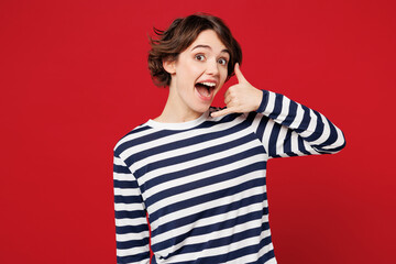 Young surprised happy woman she wears blue striped sweatshirt casual clothes doing phone gesture like says call me back look camera isolated on plain red background studio portrait. Lifestyle concept.