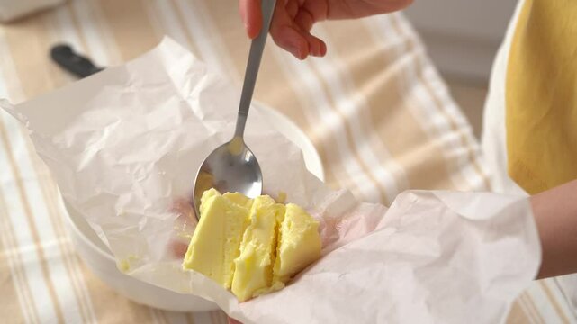 Close-up of a young woman taking spoonful of homemade yellow butter cut from parchment and putting it in bowl for further preparation at kitchen table. Concept of ingredients for dishes, cooking hobby