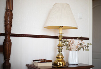 A classic brass table lamp sits on a wooden nightstand alongside a white vase with dried plants and a stack of books. The elegant setting exudes warmth, sophistication, and vintage charm.