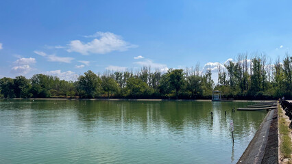 Obraz premium Calm lake with lush green trees, reflections on the water, and wooden piers under a clear blue sky, perfect for eco-tourism, relaxation, and nature-focused themes.