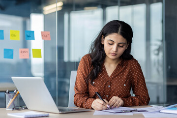 Woman working at desk in modern office, focusing on writing, planning, and paperwork. Tools such as...