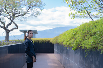 Young asian woman wearing headphones enjoys music while standing in a modern rooftop garden with lush greenery and a view of distant mountains