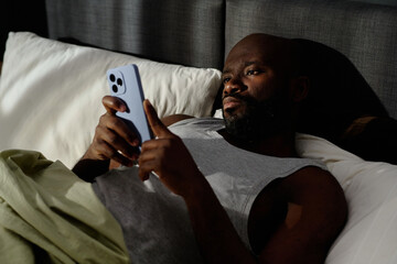High angle shot of African American man scrolling social networks while lying in bed and suffering...