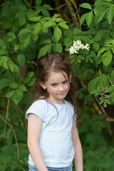 Little cute girl in t-shirt standing in front of green bushes in summer park. Healthy cheerful shy...